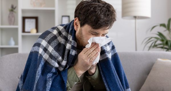 Mann, eingewickelt in einer Decke auf einem Sofa, niest in ein Taschentuch.