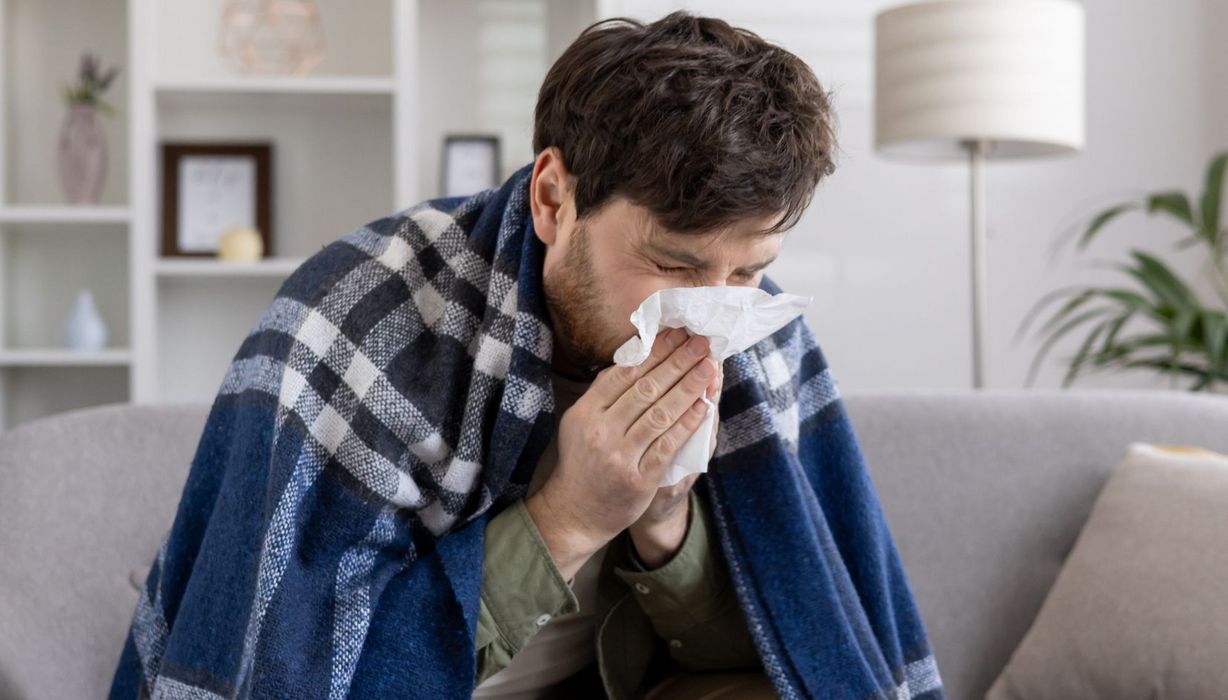 Mann, eingewickelt in einer Decke auf einem Sofa, niest in ein Taschentuch.
