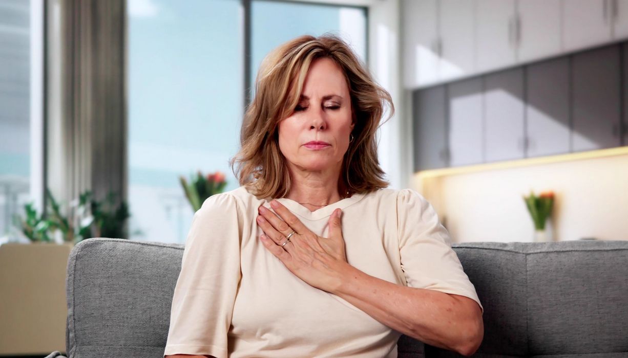 Eine Frau mittleren Alters sitzt auf einem Sofa. Sie hat geschlossene Augen und fasst mit der Hand an den tiefliegenden Halsbereich.
