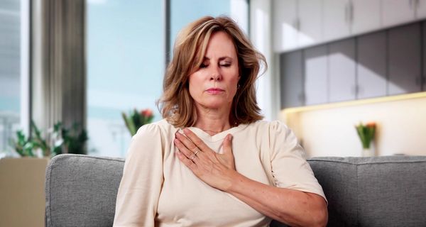 Eine Frau mittleren Alters sitzt auf einem Sofa. Sie hat geschlossene Augen und fasst mit der Hand an den tiefliegenden Halsbereich.