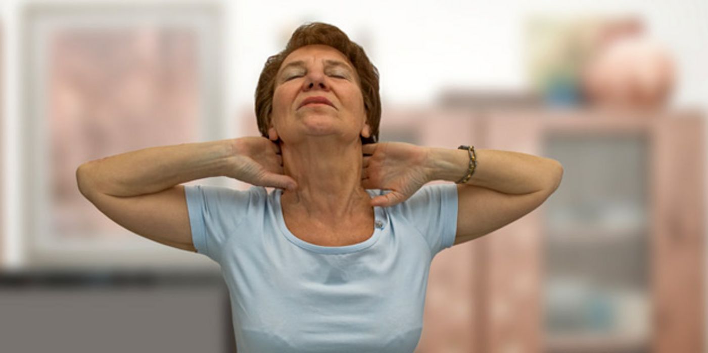 Ältere Frau sitzt am Schreibtisch und legt ihre Hände an den schmerzenden Nacken