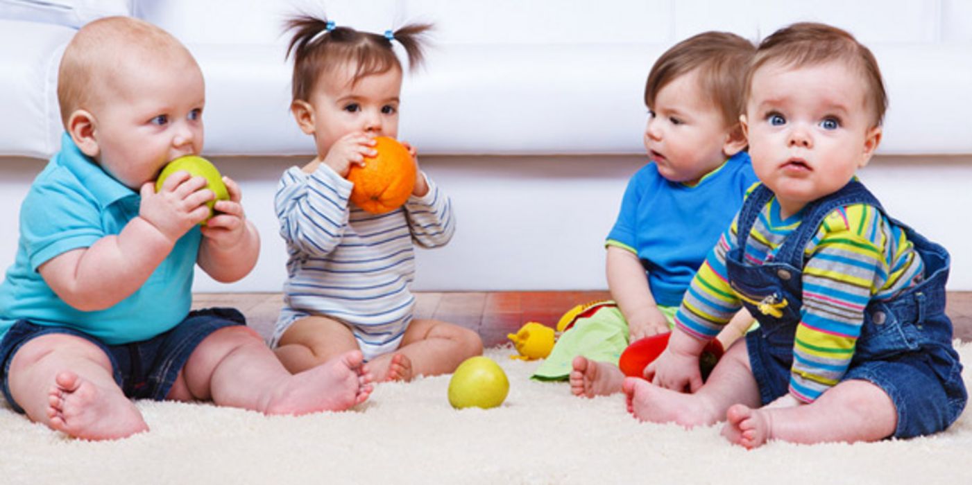 Vier Kleinkinder in farbenfroher Kleidung auf Krabbeldecke sitzend mit verschiedenen ein Mädchen beißt in Orange, ein Junge in Apfel, andere Gegenstände auf der Decke