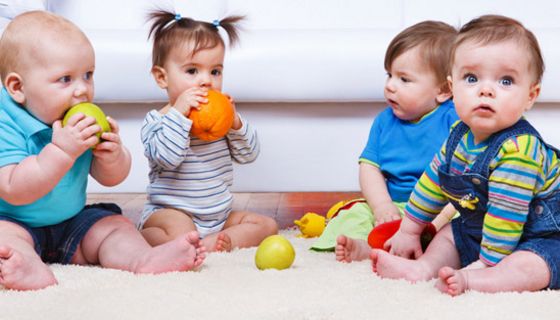 Vier Kleinkinder in farbenfroher Kleidung auf Krabbeldecke sitzend mit verschiedenen ein Mädchen beißt in Orange, ein Junge in Apfel, andere Gegenstände auf der Decke
