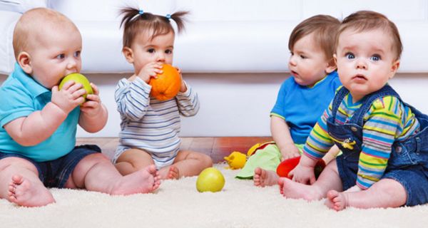 Vier Kleinkinder in farbenfroher Kleidung auf Krabbeldecke sitzend mit verschiedenen ein Mädchen beißt in Orange, ein Junge in Apfel, andere Gegenstände auf der Decke