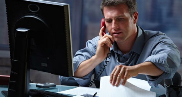 Jüngerer Mann abends im Büro am Schreibtisch mit PC, Unterlagen und Telefon am Ohr