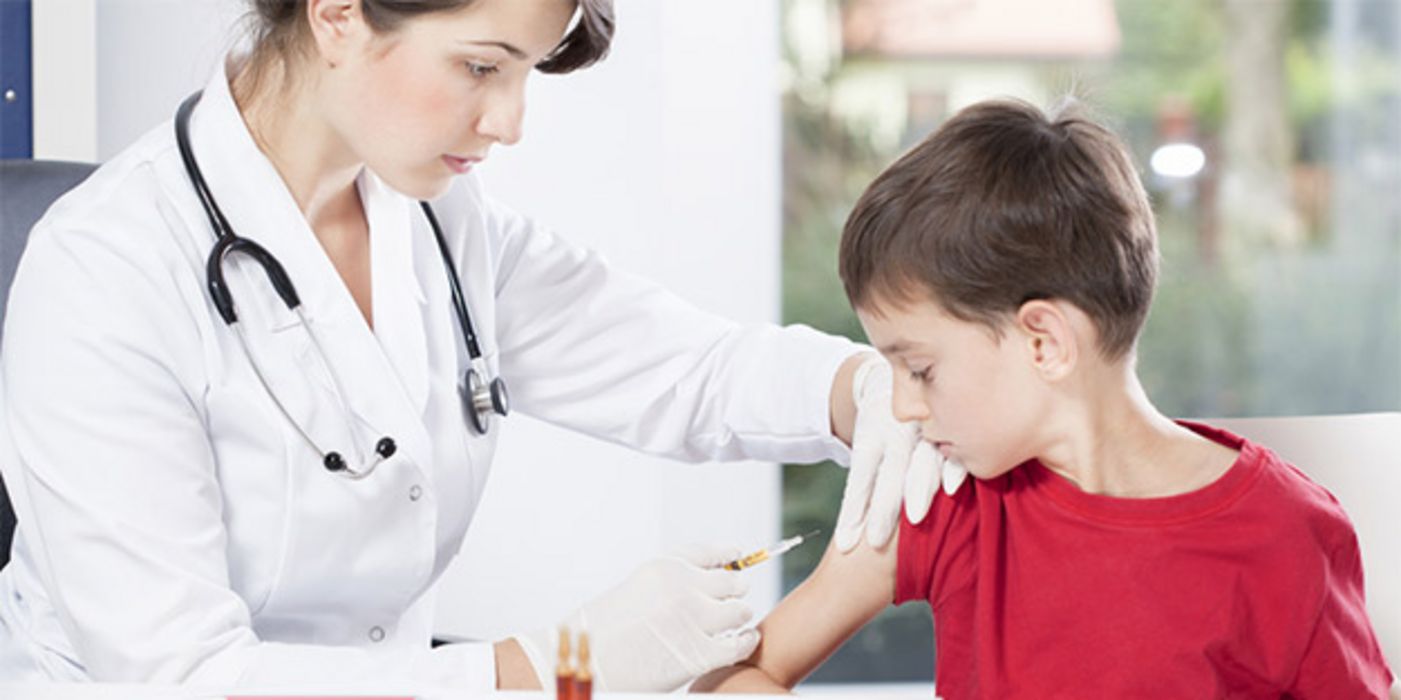 Ärztin, linker Bildrand, dunkle Haare, in den 30ern, beim Impfen eines Jungen im roten Shirt, ca. 9 Jahre alt