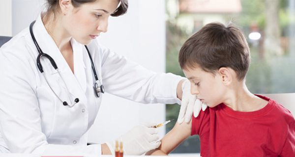 Ärztin, linker Bildrand, dunkle Haare, in den 30ern, beim Impfen eines Jungen im roten Shirt, ca. 9 Jahre alt