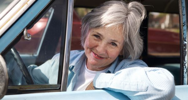 Ältere Frau, lächelt aus dem Autofenster heraus.