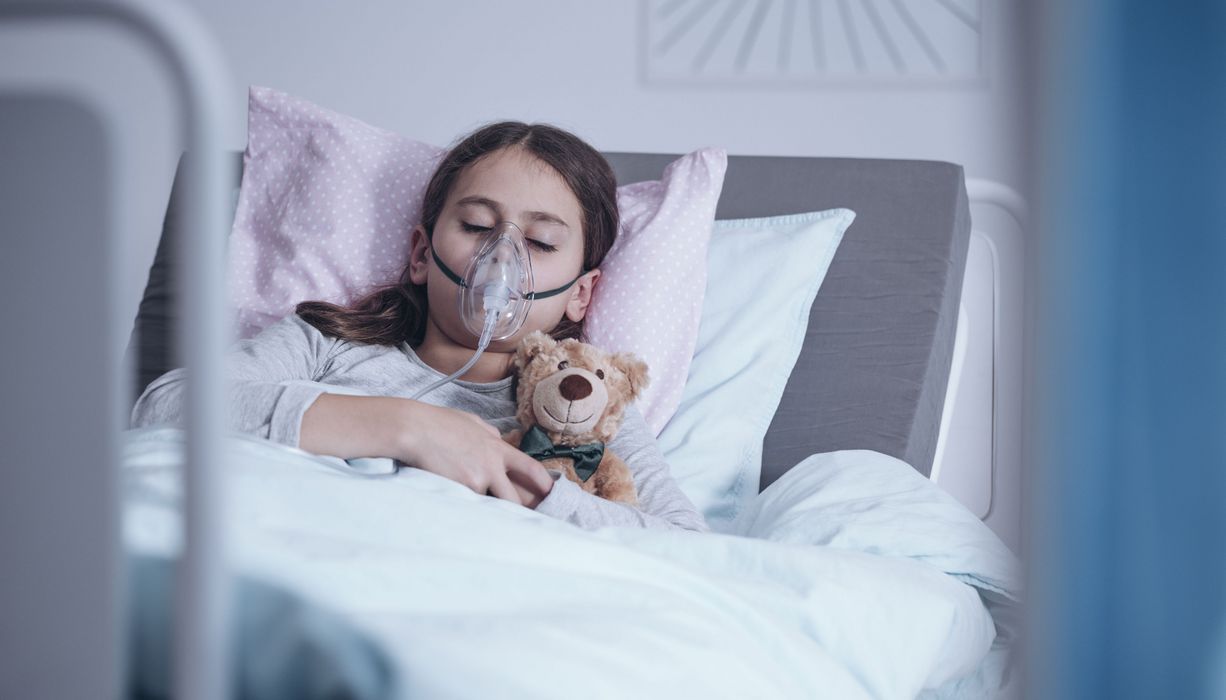 Kind liegt mit Atemmaske und Teddybär im Krankenhausbett