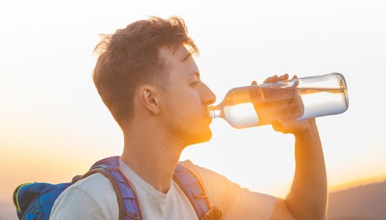 Junger Mann mit Rucksack trinkt bei Sonnenuntergang Wasser aus einer Glasflasche.