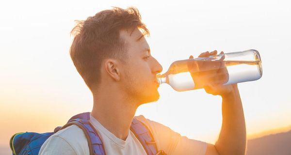 Junger Mann mit Rucksack trinkt bei Sonnenuntergang Wasser aus einer Glasflasche.