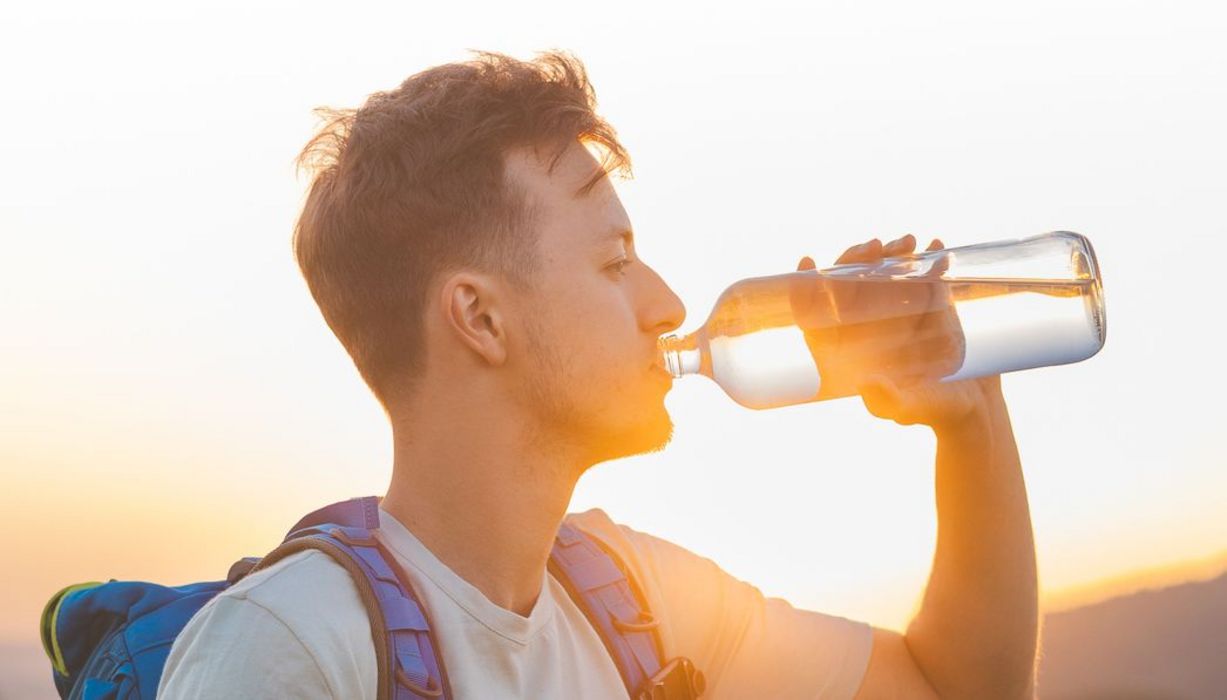 Junger Mann mit Rucksack trinkt bei Sonnenuntergang Wasser aus einer Glasflasche.