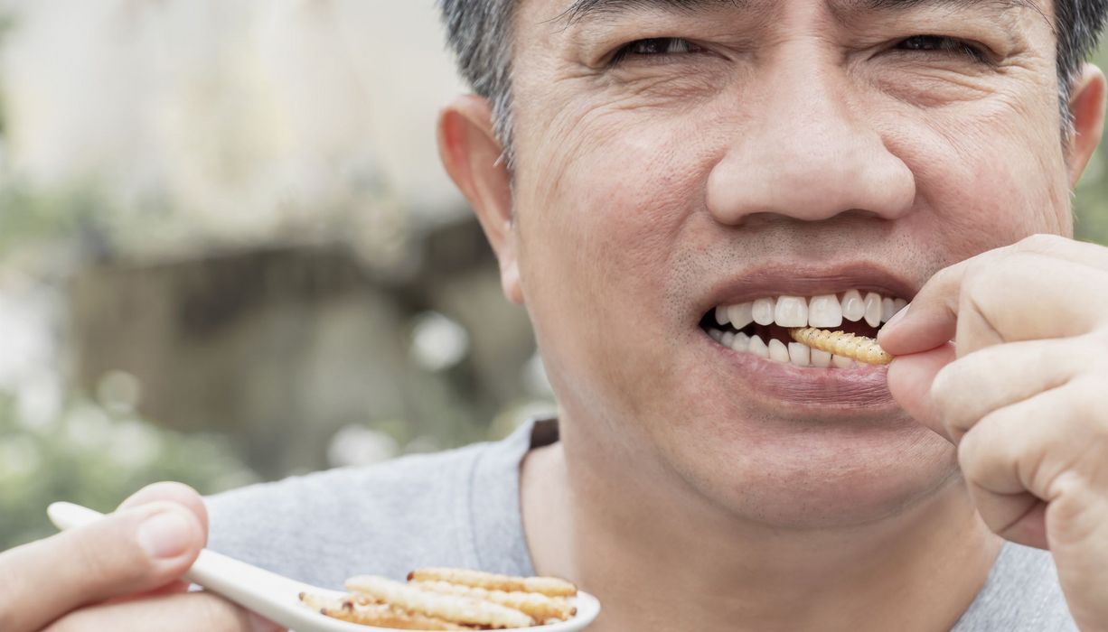Ein Mann hält einen Löffel mit Larven in seiner rechten Hand. Er steckt sich mit der anderen Hand eine Larve in den Mund und beißt darauf.