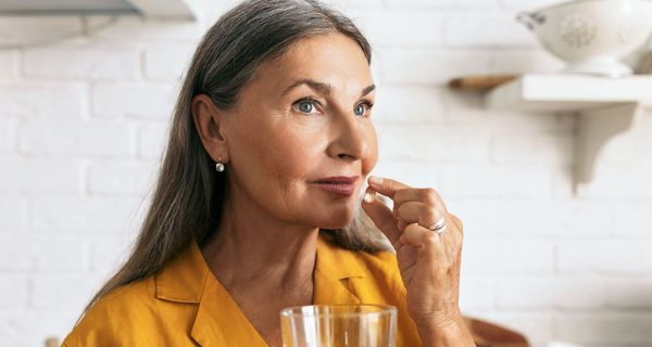 Eine ältere Dame in gelber Bluse hält eine Kapsel in der Hand und führt diese zum Mund.