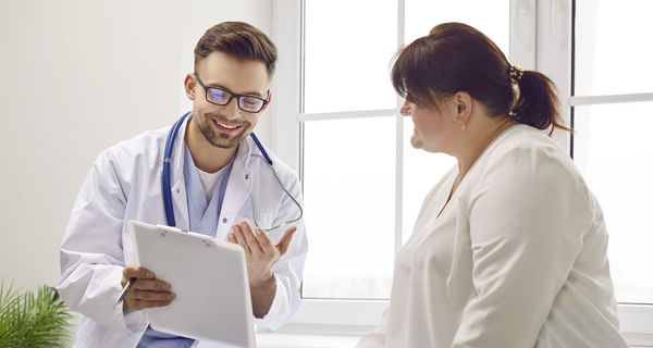 Ein Arzt zeigt lächelnd einer Frau mit Übergewicht etwas auf einem Klemmbrett.