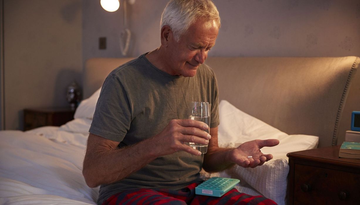 Älterer Mann, sitzt auf dem Bett und nimmt Tabletten.