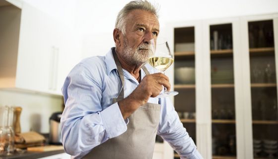 Älterer Mann steht in der Küche und trinkt Weißwein