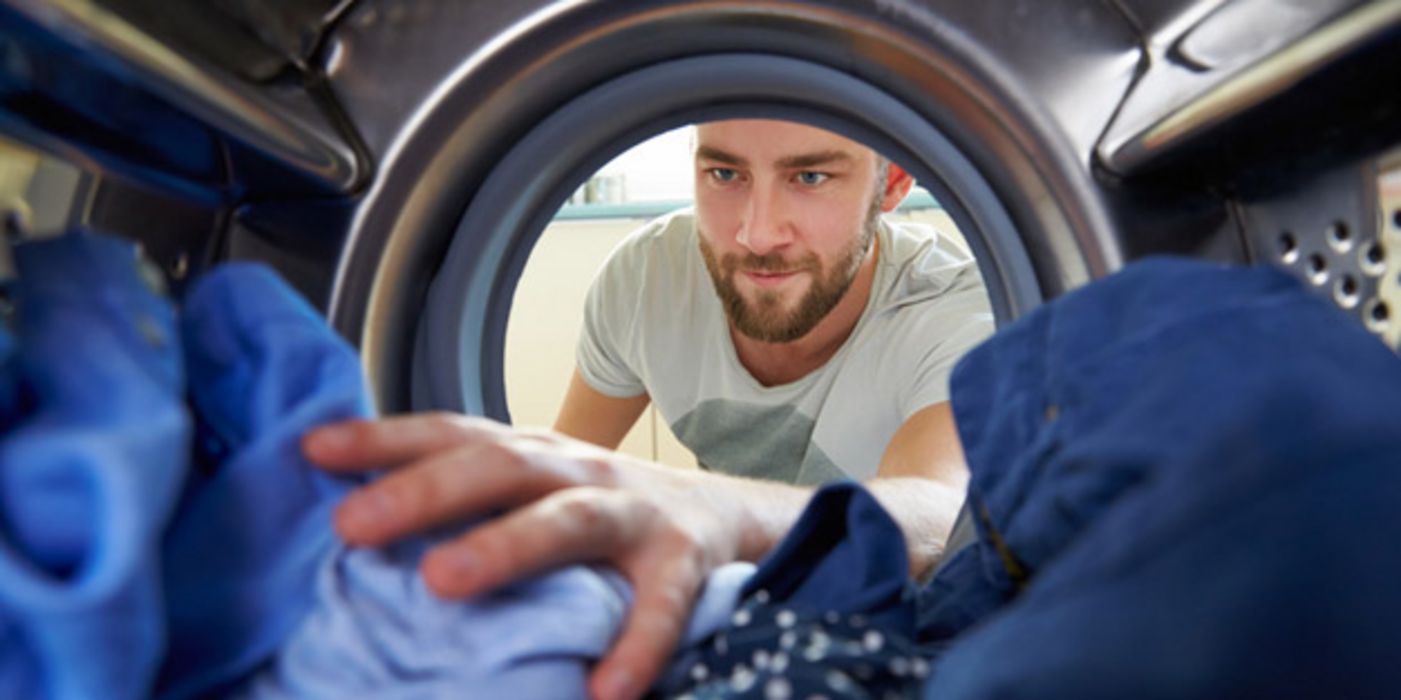 Innenansicht Wäschetrommel mit blauen und blauweißen Wäschestücken, Gesicht eines bärtigen Mannes in den 30ern in der Öffnung, ein Arm greift in die Wäsche hinein