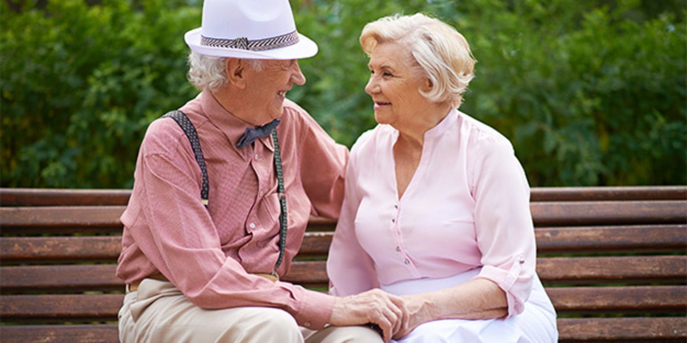 Seniorenpaar auf Bank, sich verliebt anblickend. Sommerliche, helle Kleidung, Mann hat feschen Strohhut auf