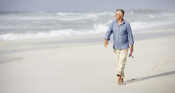 Attraktiver Senior, weißhaarig, hellblaues Hemd, weiße Hose, hochgekrempelte Hosenbeine, geht am Meer spazieren