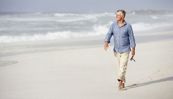 Attraktiver Senior, weißhaarig, hellblaues Hemd, weiße Hose, hochgekrempelte Hosenbeine, geht am Meer spazieren