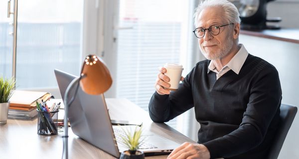 Älterer Mann sitzt mit einer Tasse Tee in der Hand vor seinem Laptop am Schreibtisch.
