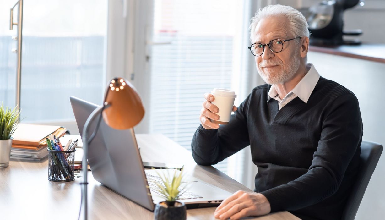 Älterer Mann sitzt mit einer Tasse Tee in der Hand vor seinem Laptop am Schreibtisch.