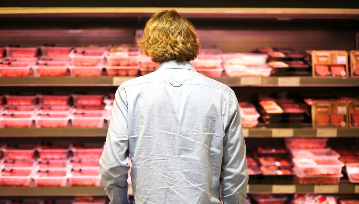 Junger Mann, steht im Supermarkt vor einem Fleischregal.