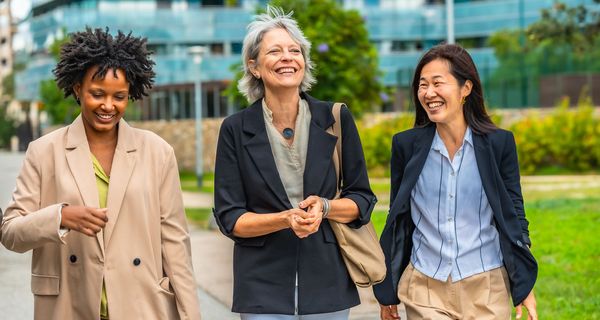Drei Frauen in geschäftlicher Kleidung gehen spazieren, im Hintergrund eine Grünfläche und ein Bürogebäude.