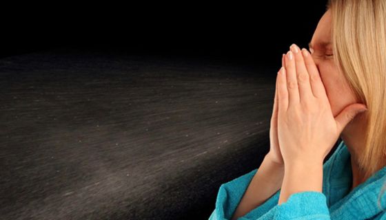 Schwarzer Hintergrund, vorne jüngere blonde Frau am Bildrand rechts im Profil, türkisfarbener Bademantel, niest in die Hände: Tröpfchennebel 