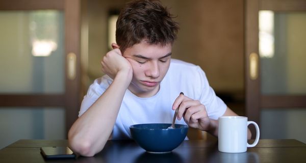 Jugendlicher, sitzt morgens sehr müde vor seinem Müsli und stochert darin herum.