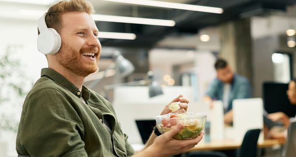 Junger Mann mit Kopfhörer isst Müsli im Büro.