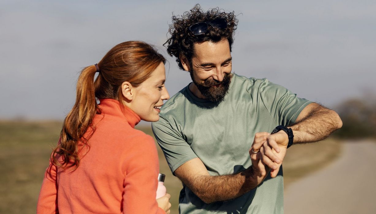Ein Paar in Sportkleidung blickt auf die Smartwatch an seinem Handgelenk, beide scheinen sich darüber zu freuen.