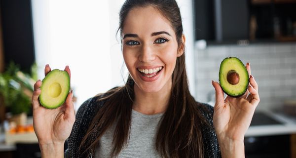 Junge, dunkelhaarige Frau hält zwei Avocadohälften in die Kamera.