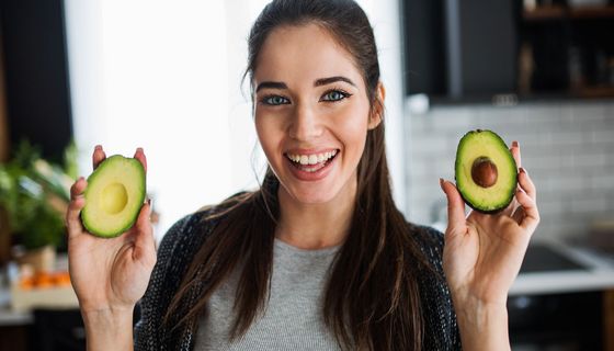 Junge, dunkelhaarige Frau hält zwei Avocadohälften in die Kamera.