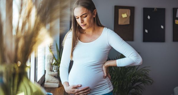 Schwangere Frau hält sich den Bauch und den Rücken. Sie blickt besorgt, der Raum hinter ihr ist dunkel.