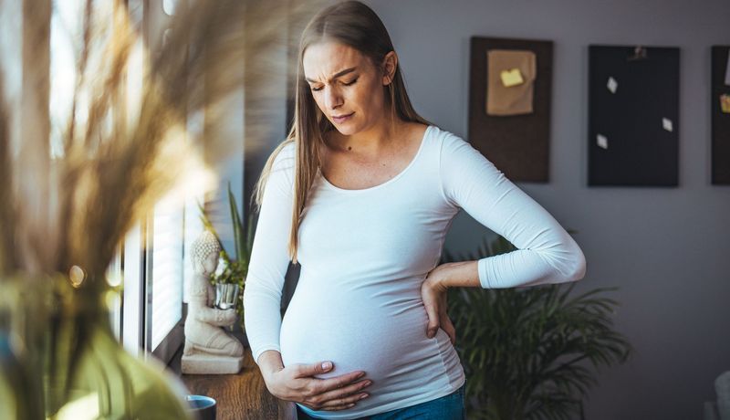 Schwangere Frau hält sich den Bauch und den Rücken. Sie blickt besorgt, der Raum hinter ihr ist dunkel.