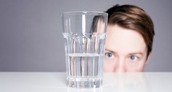 Frau lugt über eine Tischkante auf ein halb-volles Wasserglas auf dem Tisch