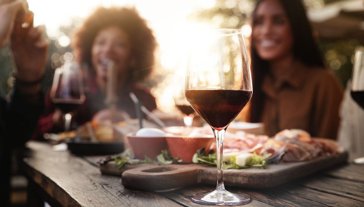 Glas Rotwein, im Hintergrund zwei Freundinnen und Essen.