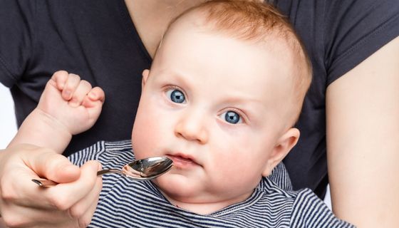 Baby bekommt einen Löffel mit einer transparenten Flüssigkeit an den Mund gehalten.