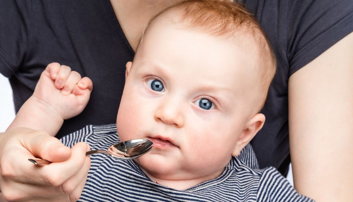 Baby bekommt einen Löffel mit einer transparenten Flüssigkeit an den Mund gehalten.