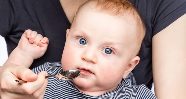 Baby bekommt einen Löffel mit einer transparenten Flüssigkeit an den Mund gehalten.