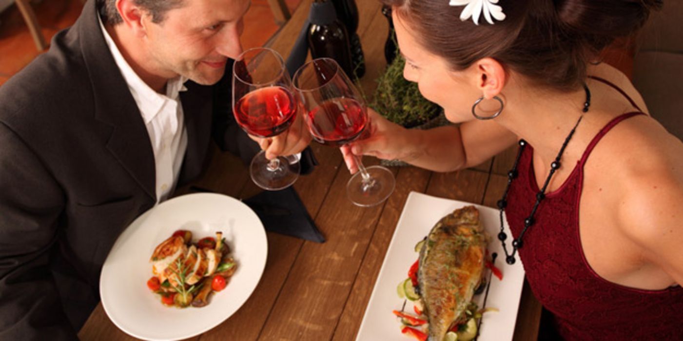 Chic gekleidetes Paar beim Dinner; sie isst Fisch; beide Partner stoßen mit einem Glas Wein an und schauen sich in die Augen