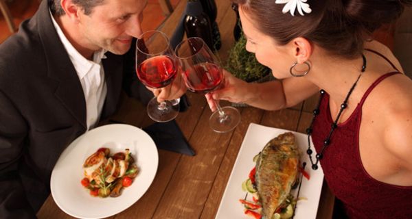 Chic gekleidetes Paar beim Dinner; sie isst Fisch; beide Partner stoßen mit einem Glas Wein an und schauen sich in die Augen
