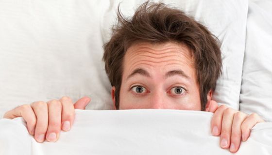 Mann schaut ängstlich unter einer Bettdecke hervor, die er bis zur Nasenspitze hochgezogen hat