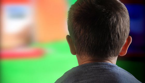 Rückenansicht eines Jungen (?) graues T-Shirt, kurze Haare, ca. 8 Jahre, bildfüllend im Hintergrund ein laufender TV-Apparat