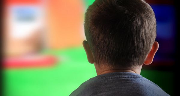 Rückenansicht eines Jungen (?) graues T-Shirt, kurze Haare, ca. 8 Jahre, bildfüllend im Hintergrund ein laufender TV-Apparat