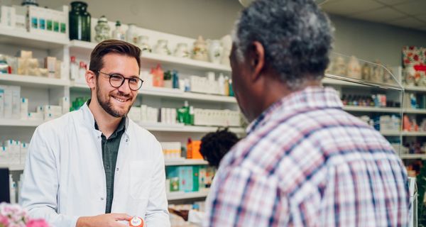 Ein lächelnder Apotheker reicht einem Kunden eine Arzneimittel. Im Hintergrund sind Regale mit weiteren Medikamenten zu sehen.