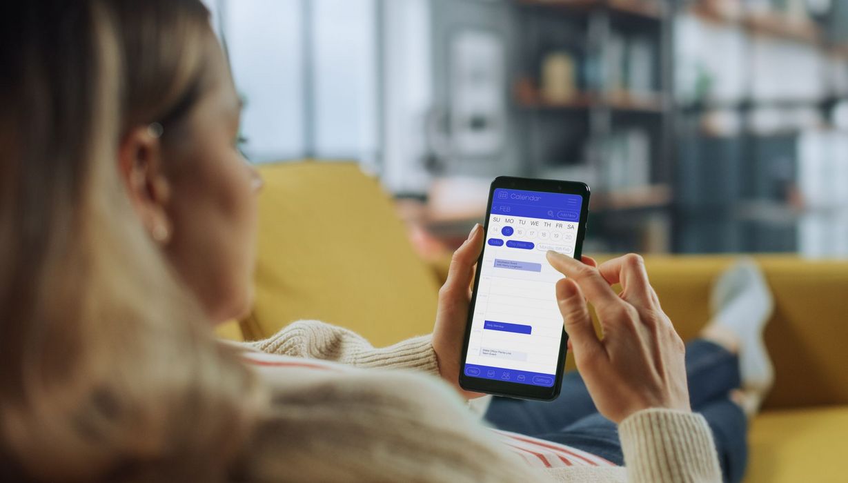Junge Frau auf dem Sofa, in der Hand ihr Smartphone mit geöffneter Kalender-App.