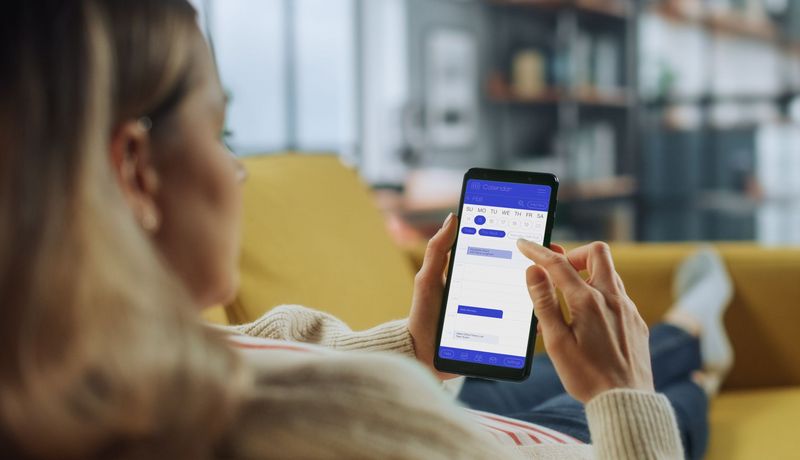 Junge Frau auf dem Sofa, in der Hand ihr Smartphone mit geöffneter Kalender-App.